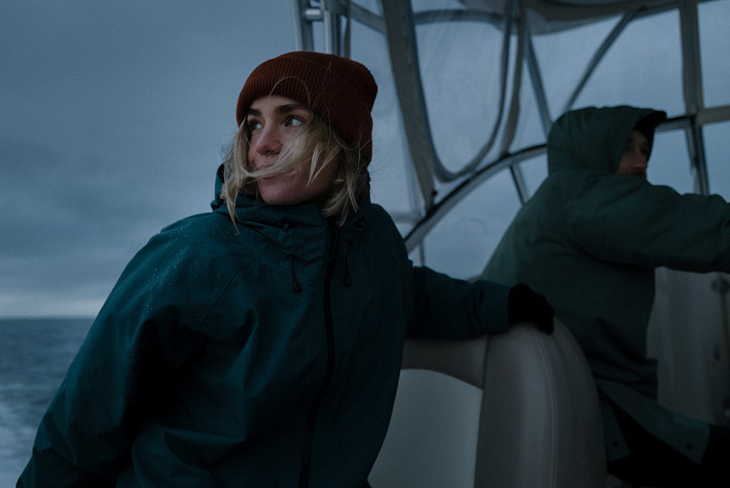 Stormy photo of woman on a boat in open water, looking over her shoulder, wearing San Poncho waterproof, breathable rain jacket in colour Dark Sea green